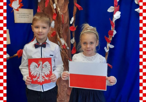 Filip i Maja w odświętnym ubraniu trzymają w rączkach godło i flagę Polski, w tle dekoracja okolicznościowa